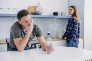 An alcoholic man drinks vodka at the kitchen table while his partner looks on worried